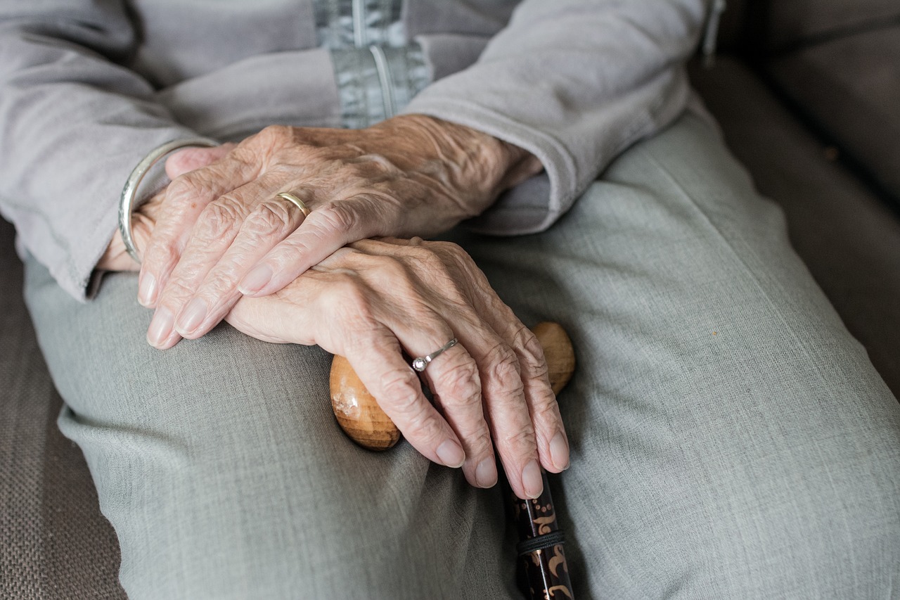 Foto de um par de mãos envelhecidas, com rugas, apoiadas no suporte de madeira de uma bengala, que está encostada no colo de uma pessoa, vestindo roupas cinzas.